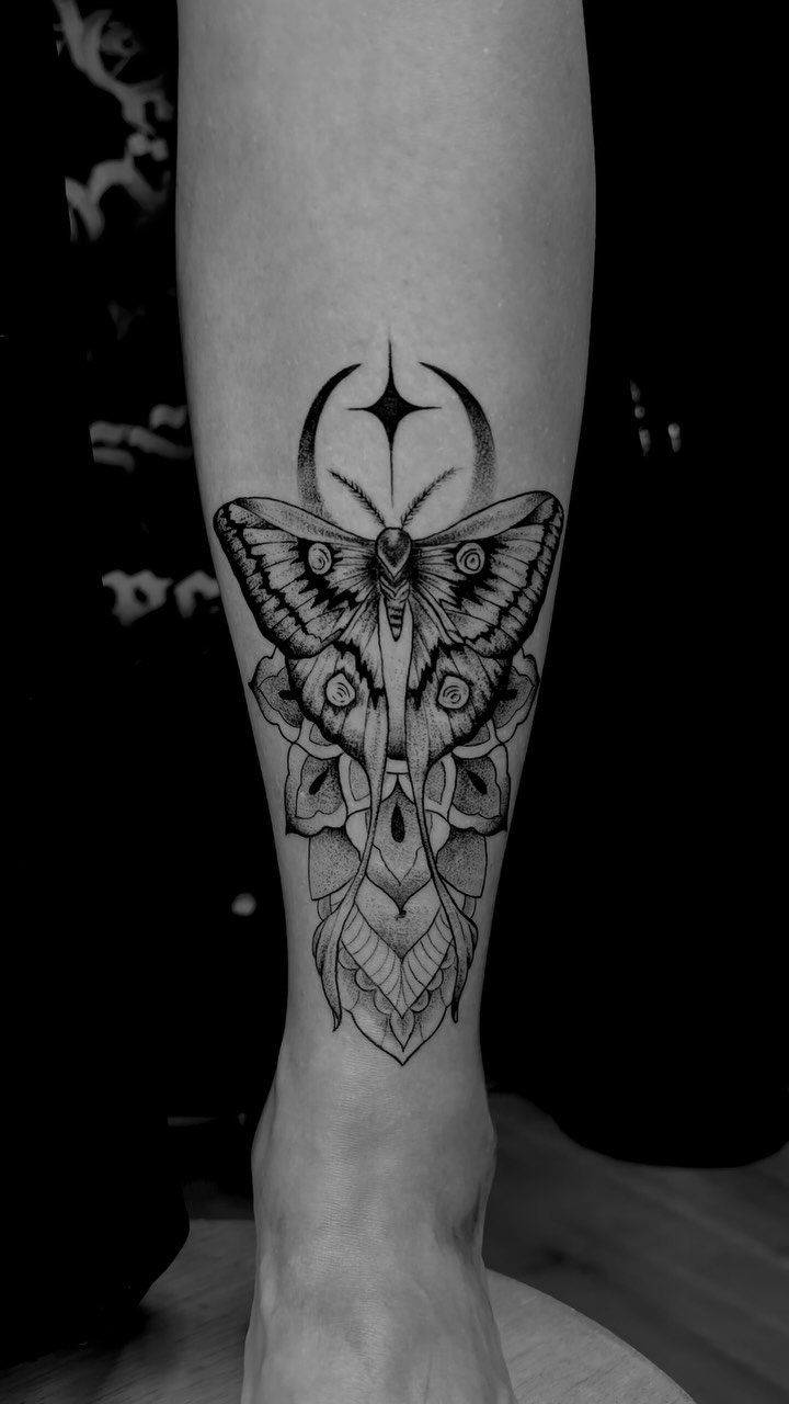 Zwart-grijze tattoo van een nachtvlinder met maansikkel en mandala op een scheenbeen.